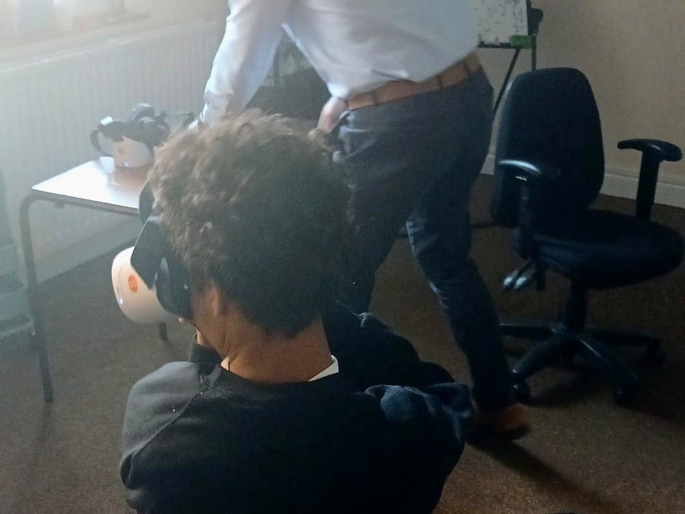 A student wearing a virtual reality headset during a lesson, with a second headset on the table beside them and a teacher working at the whiteboard behind
